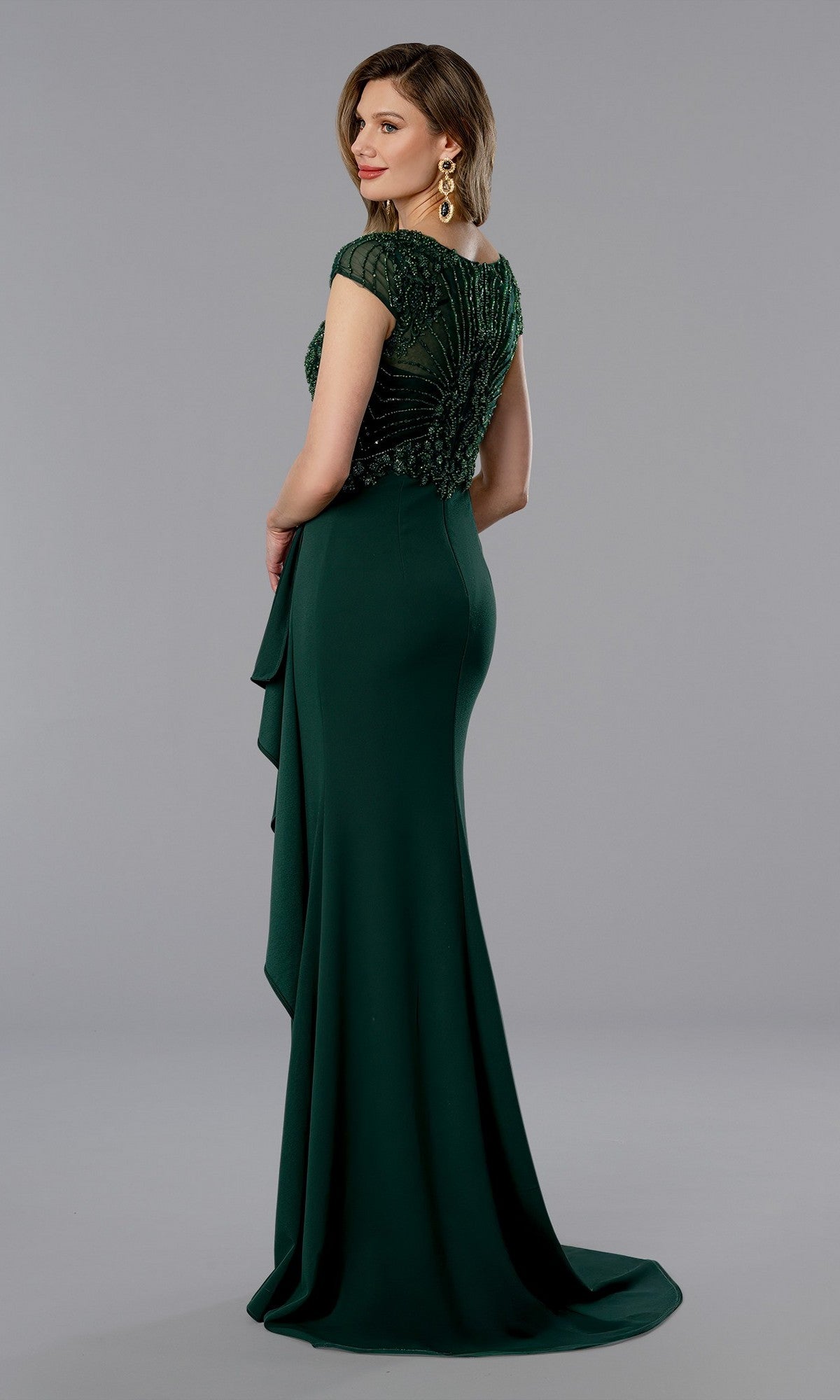Woman in a fitted, floor-length emerald green Stella Couture 25364 evening gown with beaded v-neck bodice details, standing against a plain gray background.